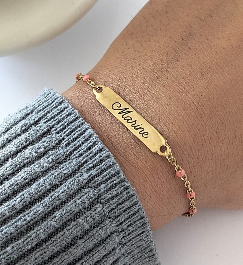 Poignet portant un bracelet personnalisé. C'est un bracelet type gourmette en acier inoxydable avec une chaine de couleur or et des perles roses. Un rectangle est gravé par un prénom ''Marine" d'une écriture cursive.