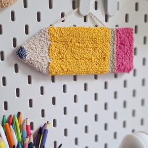 Peut inclure: Décoration murale en forme de crayon faite à la main. Le crayon présente une pointe blanche avec un accent bleu, un corps jaune, une gomme rose et une virole grise. L'œuvre est suspendue à un panneau perforé blanc. Une collection de crayons de couleur est au premier plan.