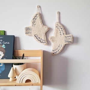 Pode incluir: Duas decorações de parede em forma de pássaro, em branco sujo, com detalhes em preto. Os pássaros são feitos de um material texturizado e têm uma argola para pendurar. Decoração de parede.