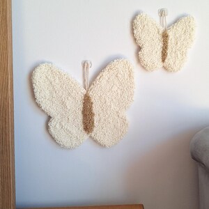 May include: Two cream-colored butterfly wall hangings with textured wings and a tan center are displayed on a white wall. The butterflies are attached with clear adhesive hooks, adding a touch of whimsy to the decor. The wall art is a charming addition to any room.