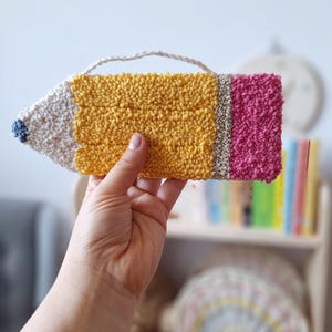 May include: A handcrafted, textured pencil-shaped wall hanging. The pencil has a yellow body, a pink eraser, and a white tip with a blue detail. A small, woven loop is attached for hanging. The pencil is held in a hand.