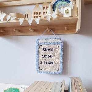 May include: A light-colored, wooden shelf displays decorative items, including miniature houses, a rainbow, and a sign that reads "Once upon a time...". Below, a row of colorful children's books is visible.