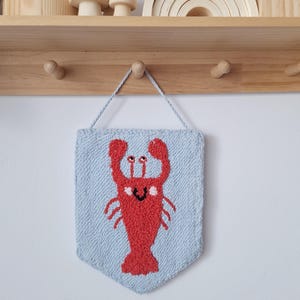 Puede incluir: Un tapiz de pared rectangular azul claro con un diseño de langosta roja. La langosta tiene una cara sonriente y está centrada en el tapiz. El tapiz está hecho de un material texturizado y cuelga de un gancho de pared de madera.