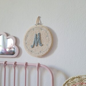 May include: A round, cream-colored embroidery hoop with the letter "M" stitched in blue yarn. The hoop is hanging on a white wall, near a pink bed frame and a cloud-shaped mirror.