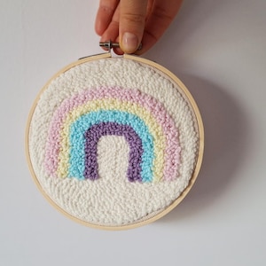 May include: A handmade, round, fiber art piece featuring a colorful rainbow design. The rainbow is composed of pastel pink, yellow, blue, and purple arches, set against a textured white background. The artwork is framed in a natural wood embroidery hoop.