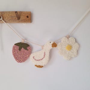 Op de afbeelding: Een decoratieve slinger met een aardbei, een gans en een bloem. De aardbei is roze met een groene bovenkant. De gans is wit met een gele snavel en poten. De bloem is wit met een geel hart. De slinger hangt aan een houten haak.