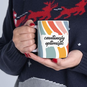 Puede incluir: Taza de cerámica blanca con un diseño de arcoíris colorido y las palabras "cautiously optimistic" en escritura negra. La taza tiene un asa blanca y la sostiene una persona que lleva un suéter azul.
