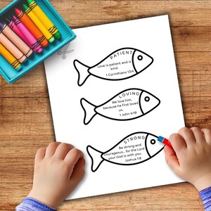 May include: A white paper with three fish-shaped illustrations, each containing a word: "PATIENT", "LOVING", and "STRONG", along with related Bible verses. A child is coloring the bottom fish with a red crayon. A teal crayon box is in the upper left.