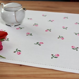 Könnte beinhalten: Weißer Tischläufer mit einem zarten rosa und grünen Blumenmuster. Der Läufer ist mit kleinen rosa Blüten und grünen Blättern bestickt. Eine weiße Teekanne und ein Tulpenstrauß liegen auf dem Tischläufer.