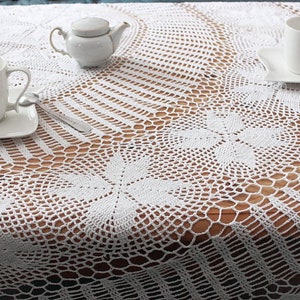 Könnte beinhalten: Weißes, aufwendig gehäkeltes Tischtuch mit floralem und geometrischem Muster. Der Tisch ist mit weißen Teetassen, Untertassen, einer Teekanne und Besteck gedeckt. Das Tischtuch hat ein zartes, offenes Gewebe.