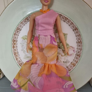 May include: A doll wearing a pink one-shoulder top and a floral print skirt with orange, pink, and yellow hues. The doll has blonde hair and a matching orange necklace. The doll is standing in front of a decorative plate.