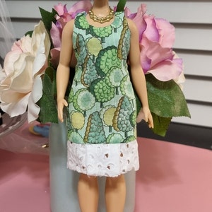 Könnte beinhalten: Eine Puppe trägt ein grün-weißes Kleid mit weißem Spitzenbesatz. Das Kleid hat ein Muster aus grünen Bäumen. Die Puppe hat blonde Haare in einem Dutt und trägt ein rotes Stirnband. Die Puppe steht auf einer rosa Oberfläche.