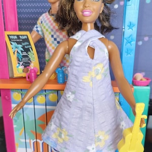 May include: A doll wearing a light purple dress with a floral pattern and holding a yellow ukulele. The doll has brown hair and brown eyes. The doll is standing in front of a colorful stand with a sign that says "Drink Up!"