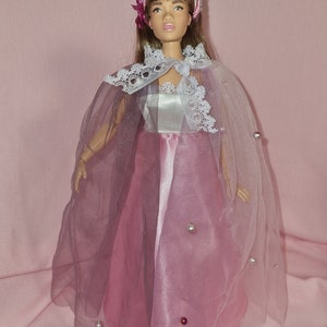 May include: A doll wearing a pink and lavender gown with a lace trim and a sheer cape. The doll has brown hair and is wearing a pink headband with a flower.