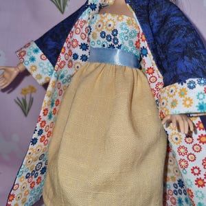 Peut inclure: Une poupée portant une robe jaune avec une ceinture bleue et une veste à imprimé floral bleu et blanc. La veste a un motif floral bleu au dos et un motif floral blanc à l'avant.