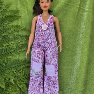 Puede incluir: Una muñeca con un mono floral morado y blanco con un acento de flor rosa. La muñeca tiene el pelo castaño y los ojos marrones.