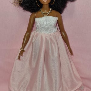 May include: A black doll with an afro hairstyle wearing a white strapless top and a pink satin skirt with a lace trim. The doll is wearing gold hoop earrings and a gold necklace.