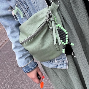 Könnte beinhalten: Eine hellgrüne Bauchtasche mit silbernem Reißverschluss und kleinem Quastenanhänger. Die Tasche ist an einem silbernen Metallverschluss befestigt. Ein Perlenanhänger mit schwarzen und grünen Perlen hängt an der Tasche.