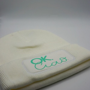 Könnte beinhalten: Cremefarbene Beanie-Mütze mit gerippter Strickstruktur. Ein rechteckiger Aufnäher auf der Vorderseite zeigt die Worte "OK. Ciao." in türkisfarbener Schrift. Die Mütze ist aus weichem Material gefertigt.