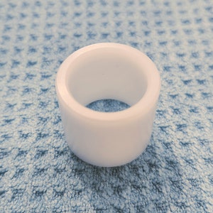 May include: A solid white, cylindrical object with a hollow center. The object is made of a smooth, opaque material and sits on a textured, light blue surface. The shape is a short, thick ring.