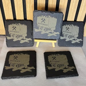 Könnte beinhalten: Ein Set aus sechs quadratischen Schieferuntersetzern. Jeder Untersetzer zeigt eine detaillierte Gravur eines mit Kohle gefüllten Bergmannswagens mit dem Wort "GLÜCKAUF" und einem Spitzhackensymbol. Die Untersetzer sind grau und schwarz.