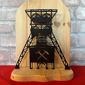 Könnte beinhalten: Schwarze Metall-Silhouette eines Bergwerksförderturms, montiert auf einer Holzplatte. Der Förderturm hat Räder, Stützbalken und eine zentrale Struktur mit gekreuzten Spitzhacken. Der Holzsockel und die Rückseite haben eine natürliche Holzmaserung.