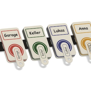 Könnte beinhalten: Vier weiße Schlüsselanhänger mit schwarzem Text und farbigen Kreisen. Die Anhänger sind mit "Garage", "Keller", "Lukas" und "Anna" beschriftet.