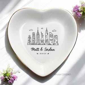 May include: White heart-shaped ceramic dish with a gold rim. The dish features a black and white illustration of the New York City skyline with the text "Matt & Jordan 30.01.25".