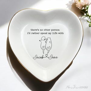 Puede incluir: Plato de cerámica blanco en forma de corazón con borde dorado. El plato presenta una ilustración en blanco y negro de dos nutrias que se toman de la mano con el texto "there's no otter person i'd rather spend my life with" y los nombres "Jacob Jane" debajo de las nutrias.