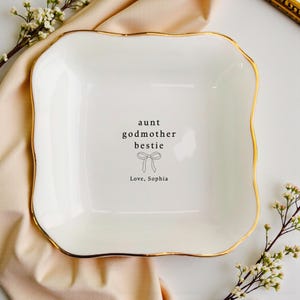 Puede incluir: Un plato cuadrado de cerámica blanca con borde dorado. El plato tiene las palabras "aunt godmother bestie" y "Love, Sophia" impresas, junto con un gráfico de lazo. El plato está rodeado de flores blancas y una tela color melocotón.