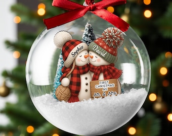 May include: A clear glass Christmas ornament with a red ribbon. Inside, two snowman figures are depicted, wearing hats and scarves. The ornament has the names "Sophie" and "David" on the snowmen, and a sign that reads "First Christmas as Mr. & Mrs."
