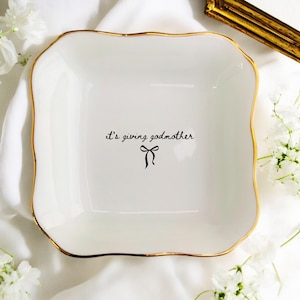 May include: White square ceramic trinket dish with a gold rim and the phrase "it's giving godmother" in black script, along with a black bow graphic. The dish has a wavy edge.