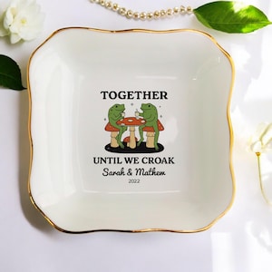 May include: A white ceramic trinket dish with a gold rim. The dish features a whimsical illustration of two green frogs sitting at a mushroom table, with the text "TOGETHER UNTIL WE CROAK" and names below.