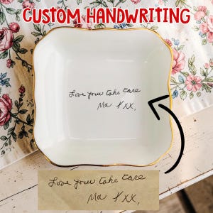 Peut inclure: Un vide-poches carré blanc avec un bord doré et des bords festonnés. Le plat présente un texte manuscrit à l'encre noire qui dit "Love you take care Ma XXX,". Les mots "CUSTOM HANDWRITING" sont en rouge en haut.