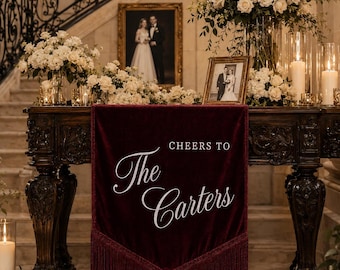 May include: A deep burgundy velvet banner with fringe trim, featuring the white text "CHEERS TO The Carters" draped over a dark ornate wooden table. The table is decorated with large white floral arrangements and tall glass pillar candles.