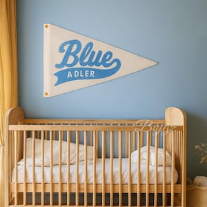 K&ouml;nnte beinhalten: Ein cremefarbener Wimpel mit dem Wort "Blue" in blauer Schrift und "ADLER" in blauer serifenloser Schrift. Der Wimpel h&auml;ngt an einer hellblauen Wand &uuml;ber einem Holzbettchen.