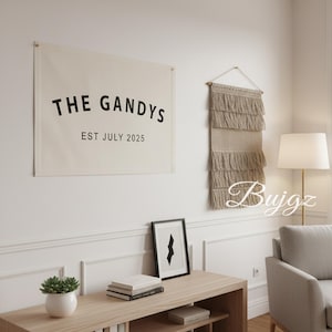 Puede incluir: Un salón de tonos neutros con un tapiz de pared de lona que dice "THE GANDYS EST JULY 2025". A su lado cuelga un tapiz de pared tejido con flecos. Una impresión en blanco y negro enmarcada y una suculenta en una maceta blanca se encuentran en una consola de madera.