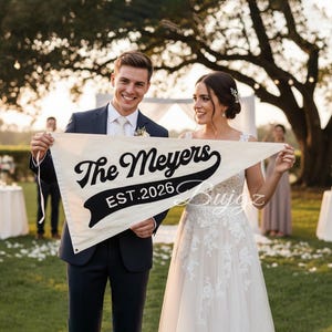 May include: A cream pennant banner with the text "The Meyers EST. 2026" in black script and a black banner. The banner is held by a smiling couple, the man in a navy suit and the woman in a white wedding dress, at an outdoor wedding.