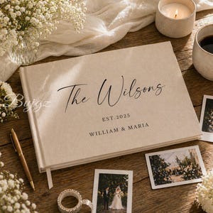 Op de afbeelding: Een rechthoekig fotoalbum met een beige stoffen kaft met de zwarte opdruk The Wilsons, EST. 2025 en WILLIAM & MARIA. Het album ligt op een houten ondergrond naast een gouden pen, een rolletje tape en twee kleine foto's.