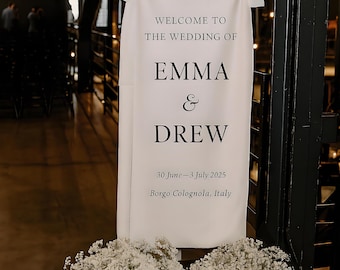 Może przedstawiać: Biały znak weselny z czarnym tekstem "WELCOME TO THE WEDDING OF EMMA & DREW" i datą "30 czerwca-3 lipca 2025". Lokalizacja to "Borgo Colognola, Włochy". Znak jest ozdobiony białymi kwiatami.