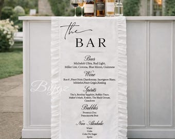 Op de afbeelding: Een witte tafelloper voor de bar met een gerimpelde rand, met de tekst "The BAR" en een menu met bier, wijn, sterke drank, bubbels en alcoholvrije dranken. Wijnglazen en flessen op de bar.