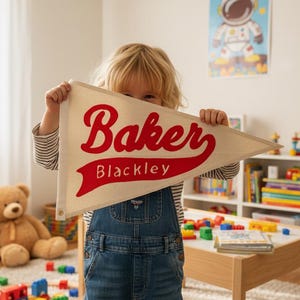 May include: A cream-colored pennant flag with the name "Baker" in red script and "Blackley" in a smaller font. The flag is held by a child in a room with toys and a teddy bear.