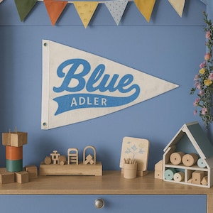 K&ouml;nnte beinhalten: Ein cremefarbenes Wandbanner mit dem Wort "Blue" in Schreibschrift und dem Namen "Adler" darunter. Das Banner h&auml;ngt &uuml;ber einem Holzregal mit Spielzeug und Dekorationsartikeln. Eine Girlande aus bunten Wimpeln h&auml;ngt dar&uuml;ber.