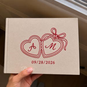 May include: A rectangular beige fabric-covered book featuring red embroidered twin heart lockets tied with a ribbon. The letters A and M are stitched inside the hearts, with the date 09/28/2026 embroidered in red below the design.