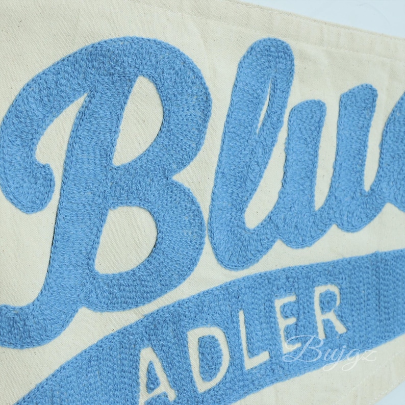 K&ouml;nnte beinhalten: Cremefarbener Wimpel mit dem Wort "Blue" in gro&szlig;er, hellblauer gestickter Schrift. Darunter ist das Wort "Adler" in der gleichen hellblauen Farbe gestickt. Der Wimpel besteht aus einem leinwandartigen Material.