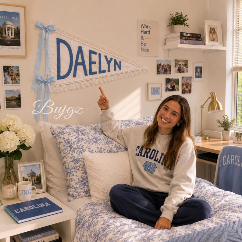 May include: A white triangular fabric pennant featuring the name DAELYN in bold blue letters, accented with light blue ribbons and white pom-pom trim. The pennant is displayed on a wall above a bed with blue and white floral bedding.