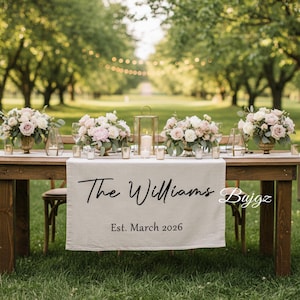 May include: A wooden table set for a wedding reception, adorned with floral arrangements in gold vases, candles, and glassware. A linen table runner displays the text "The Williams" and "Est. March 2026". The scene is set outdoors with trees and string lights.