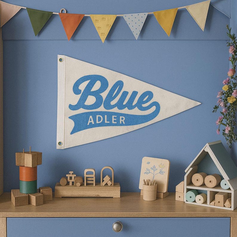 K&ouml;nnte beinhalten: Ein cremefarbenes Wandbanner mit dem Wort "Blue" in Schreibschrift und dem Namen "Adler" darunter. Das Banner h&auml;ngt &uuml;ber einem Holzregal mit Spielzeug und Dekorationsartikeln. Eine Girlande aus bunten Wimpeln h&auml;ngt dar&uuml;ber.