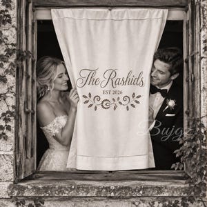 May include: A rectangular off-white fabric panel featuring the text The Rashids, EST 2026, and a decorative leaf motif. The fabric has a hemmed border with visible stitching and is positioned between a person in a wedding dress and a person in a tuxedo.