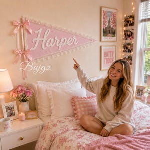 Op de afbeelding: Een grote driehoekige roze vilt wandwimpel met de naam Harper in witte schrijfletters, versierd met witte kanten randen en twee roze satijnen strikken aan de linkerkant, bevestigd aan een muur boven een bed met bloemrijk beddengoed.